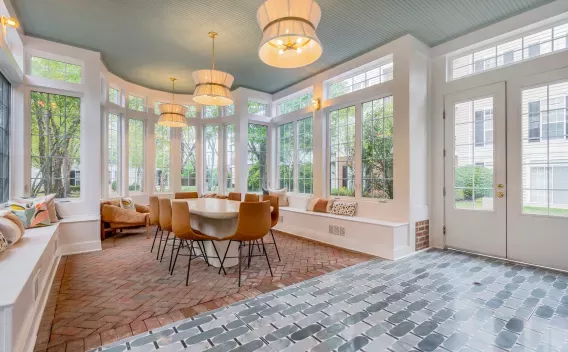 Bright, modern dining room with large windows, light blue ceiling, a long table with brown chairs, and mixed brick and tile floors.