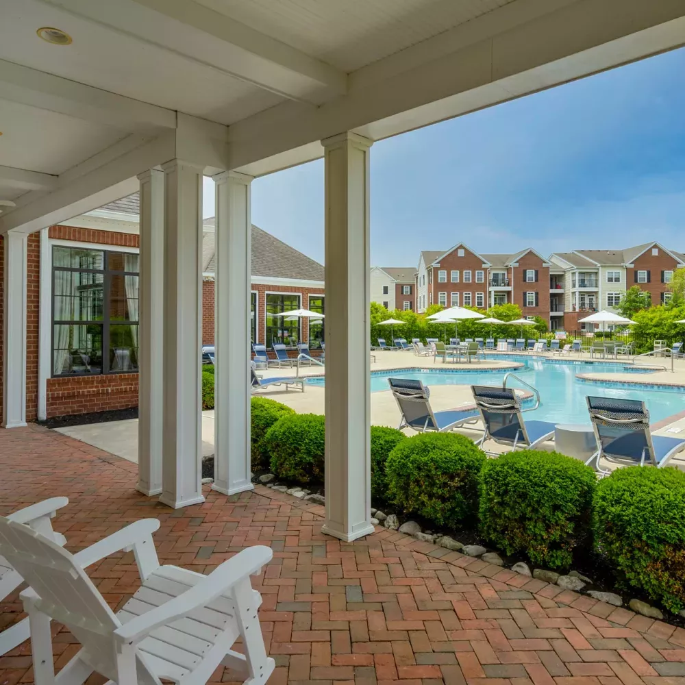 Covered patio with white Adirondack chairs overlooks a large outdoor swimming pool surrounded by lounge chairs and apartment buildings.