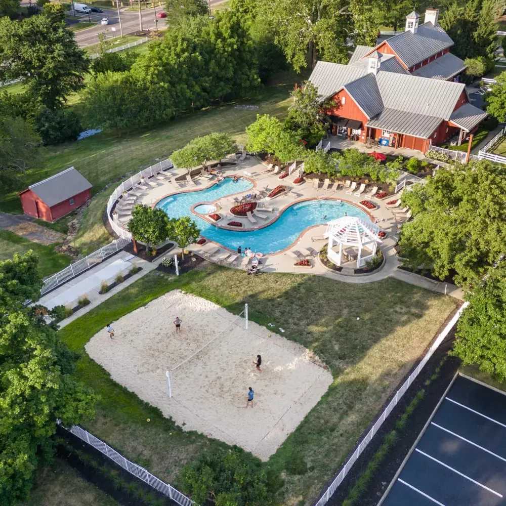 Aerial view of a recreational area with a red clubhouse, blue pool, and sand volleyball court with people playing.