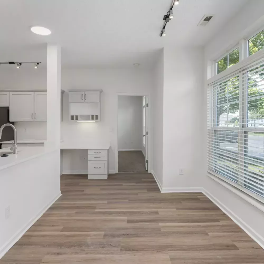 Modern apartment interior featuring a white kitchen, light wood flooring, a built-in desk, and a large window with blinds.