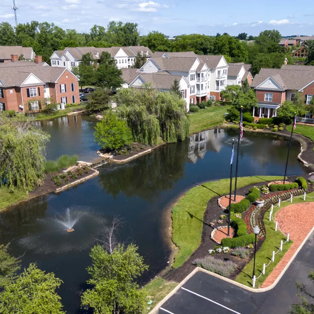Aerial photograph of a residential community with brick buildings, a pond with a fountain, green trees, and an island with flags.