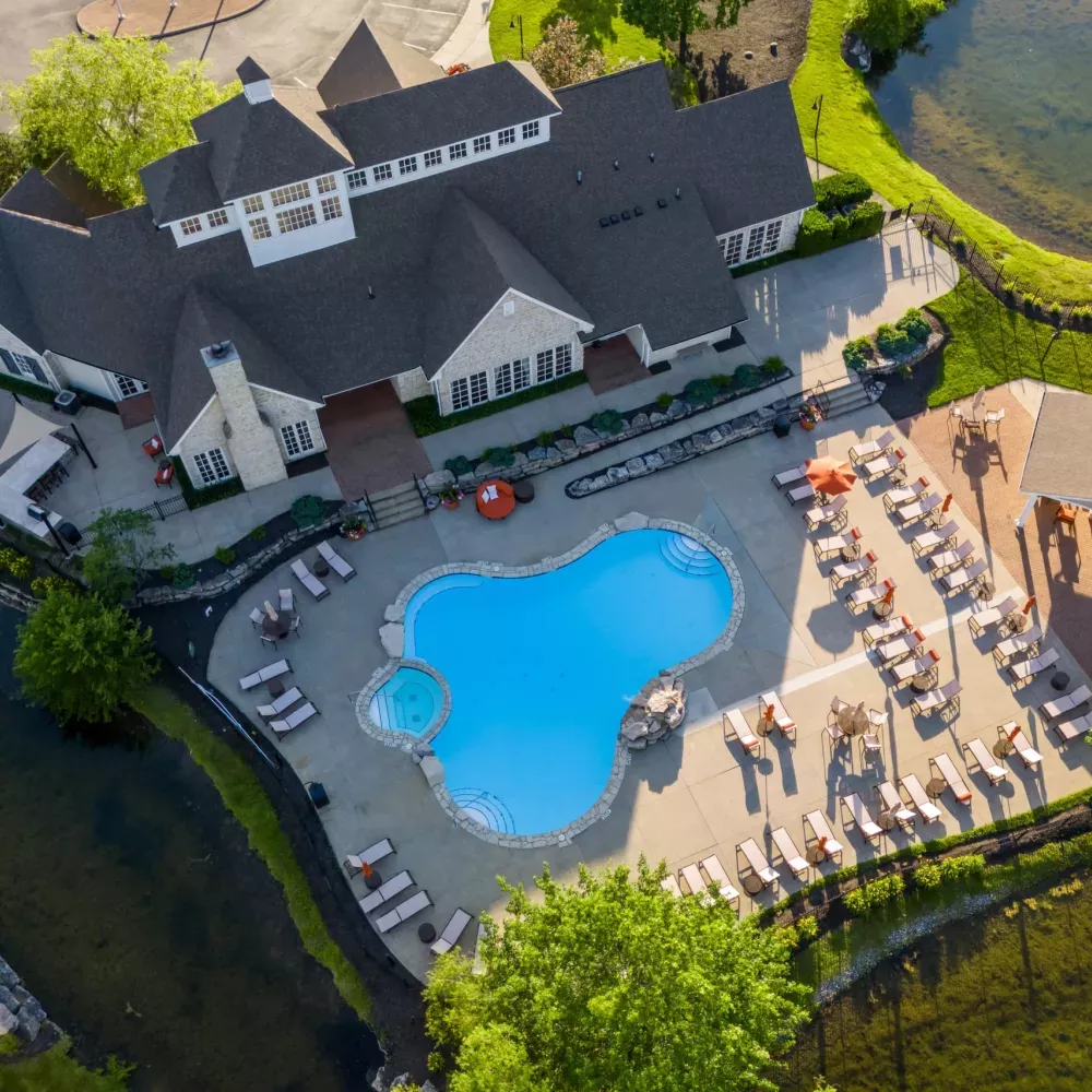 Aerial view of a clubhouse with a freeform swimming pool surrounded by lounge chairs, green lawns, trees, and two ponds.