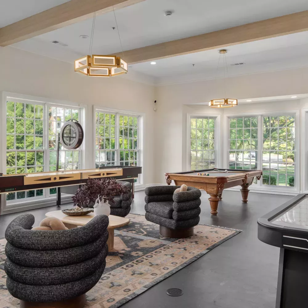 Bright recreation room with pool table, foosball, air hockey, and lounge seating by large windows.