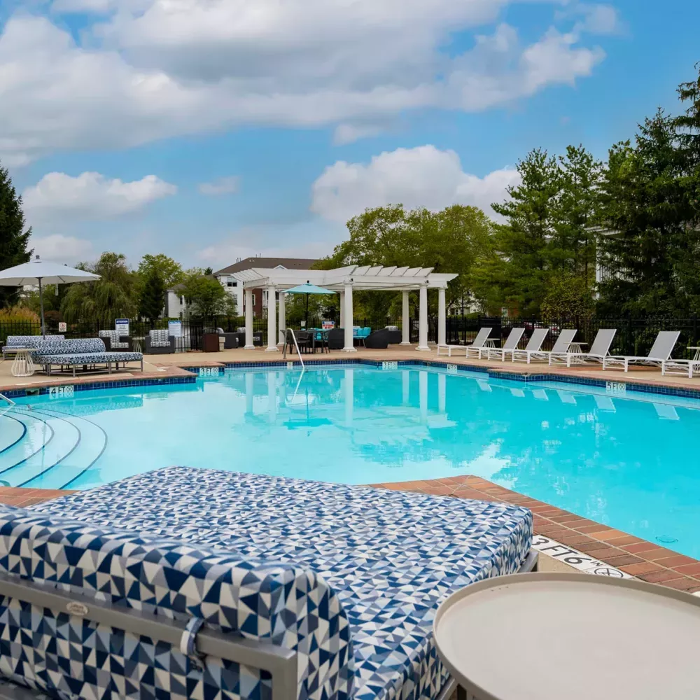 Sunny outdoor swimming pool with blue water, surrounded by lounge chairs and green trees under a partly cloudy sky.