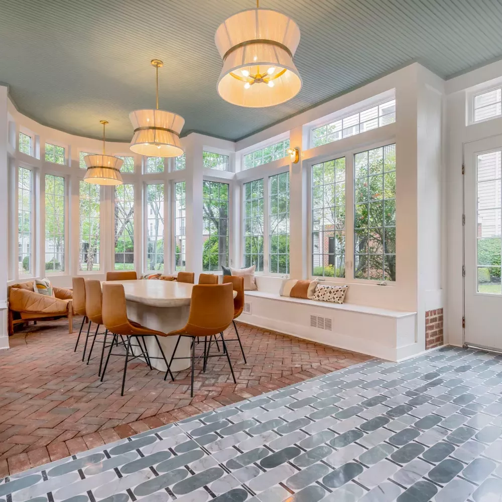 Bright, modern dining room with large windows, light blue ceiling, a long table with brown chairs, and mixed brick and tile floors.