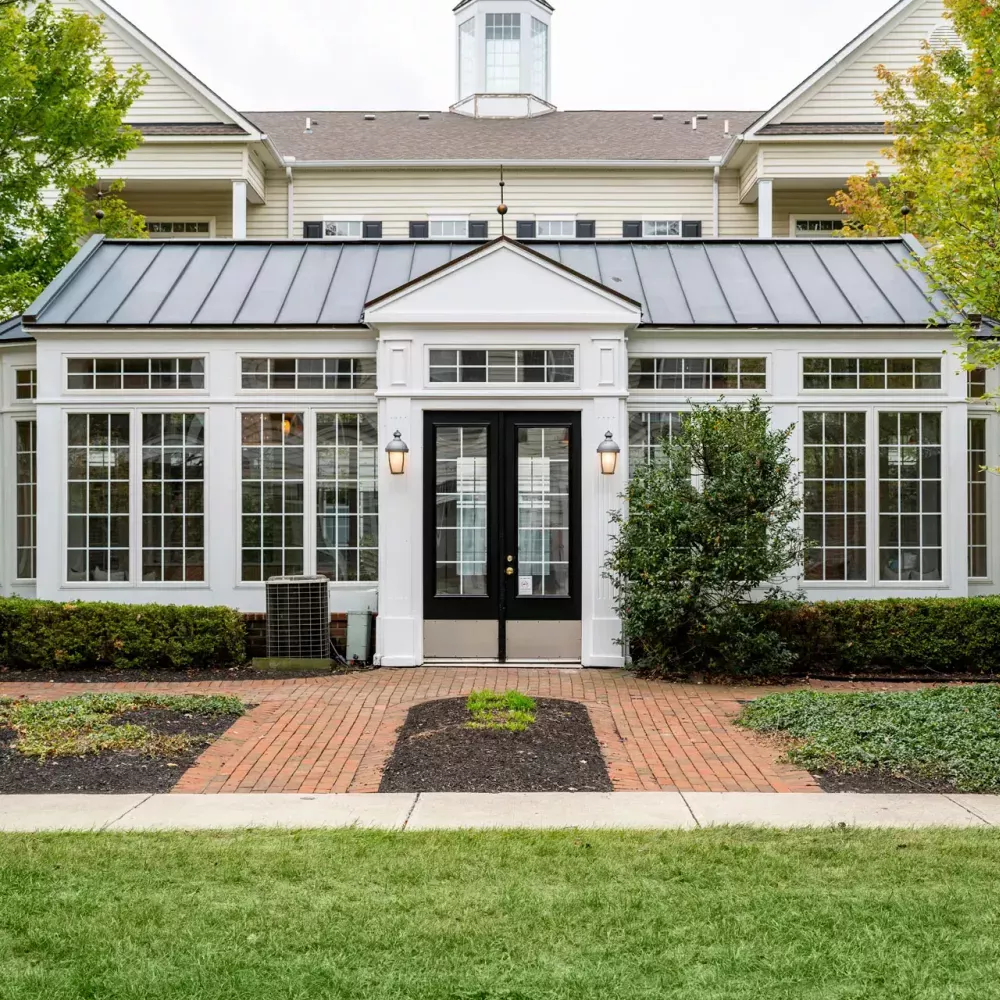 White sunroom building with a dark roof, black double doors, and many windows, surrounded by green grass and fall trees.