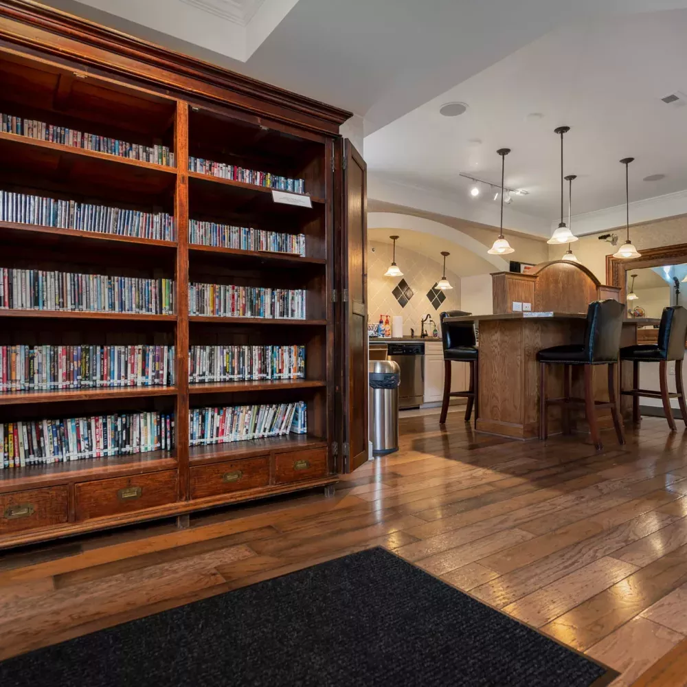 Tall wooden bookshelf filled with books and media, next to a modern bar with stools and pendant lights on a gleaming wood floor.