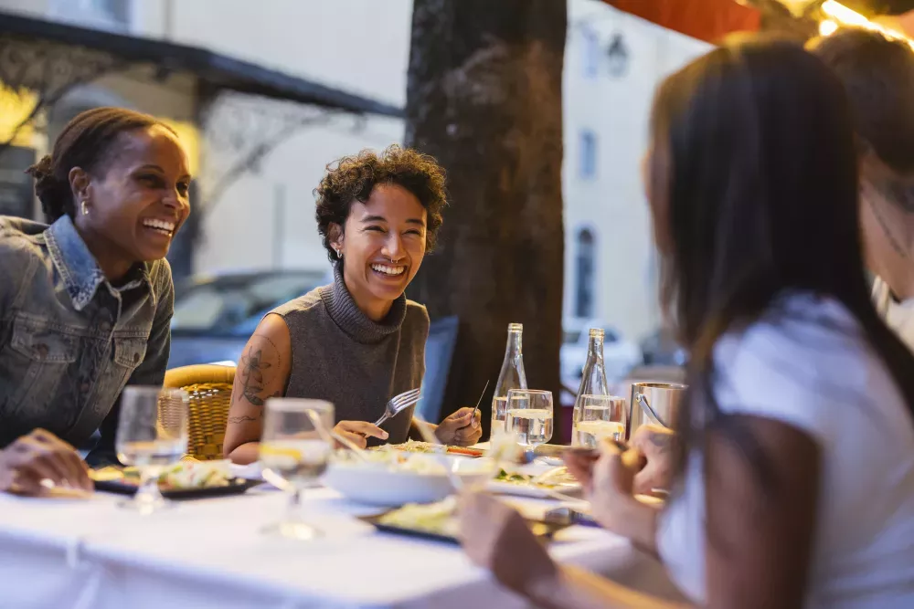 Three women laughing and sharing a meal at an outdoor restaurant table at night.