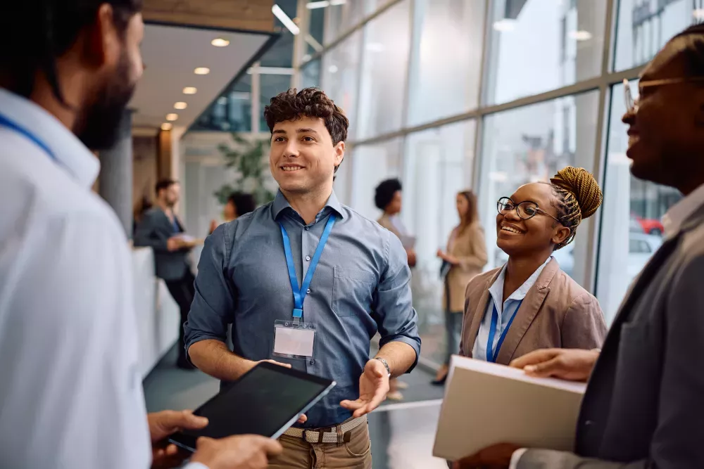 Three diverse professionals with ID badges smile and chat in a brightly lit modern office setting.